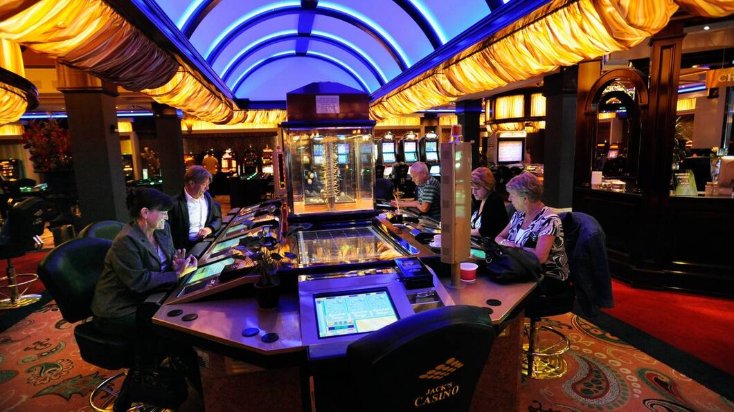 Foto van een binnenruimte in een casino, met in het middel een tafel waaraan 5 mensen zitten te gokken op een beeldschderm.